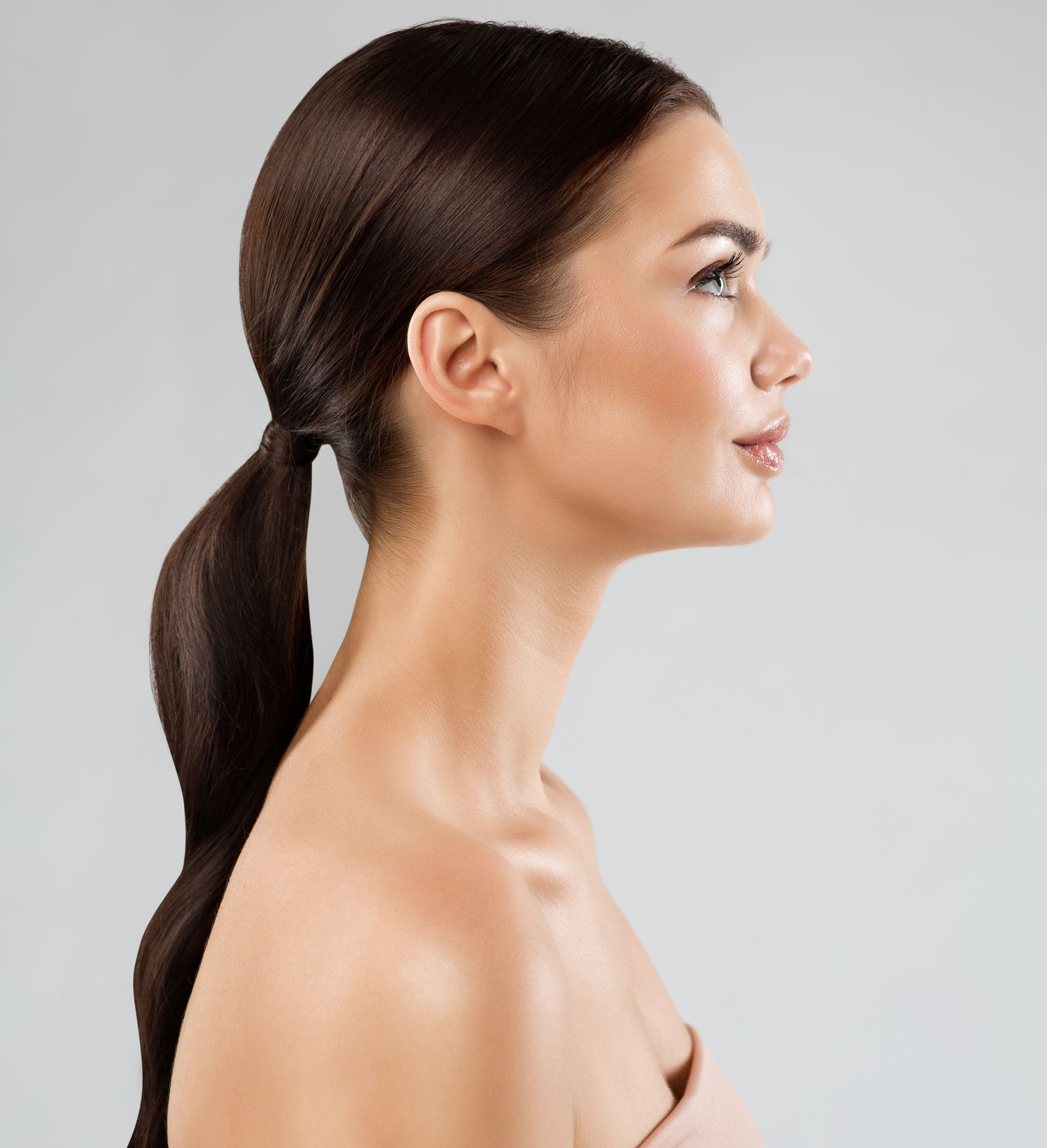Woman in profile, wearing a ponytail and a strapless top, looking to the right against a gray background.