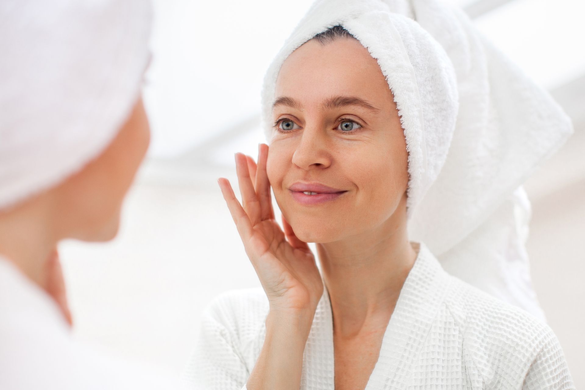 Woman in a white robe gently applying skincare cream while looking at her reflection in a mirror