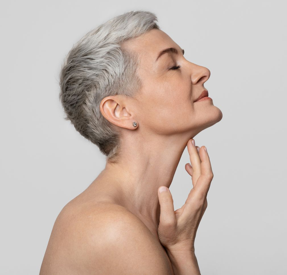 Woman with short gray hair touching her neck, eyes closed, against a gray background.
