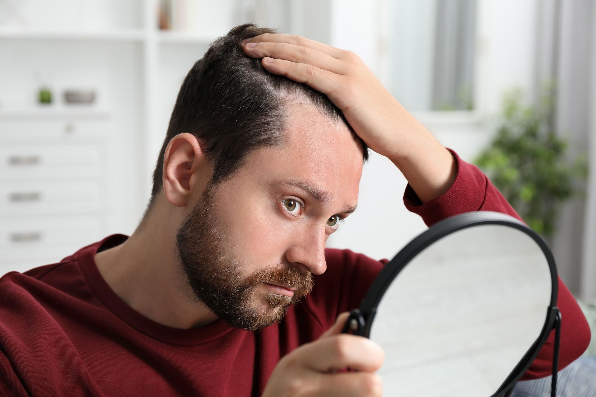 Man examining his scalp in a handheld mirror, concerned expression, indoor setting.