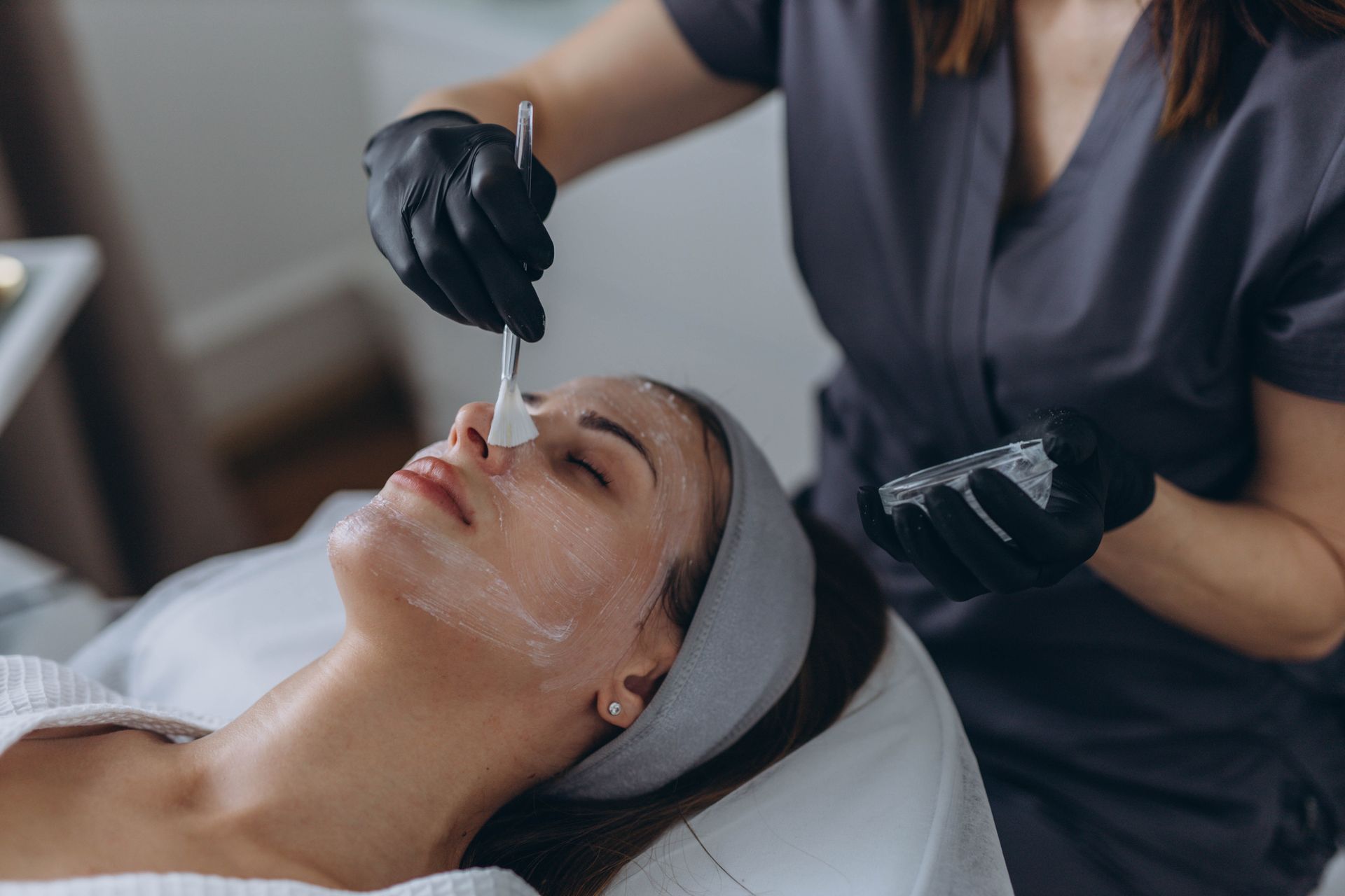 Person receiving facial treatment; esthetician applying product with a brush.