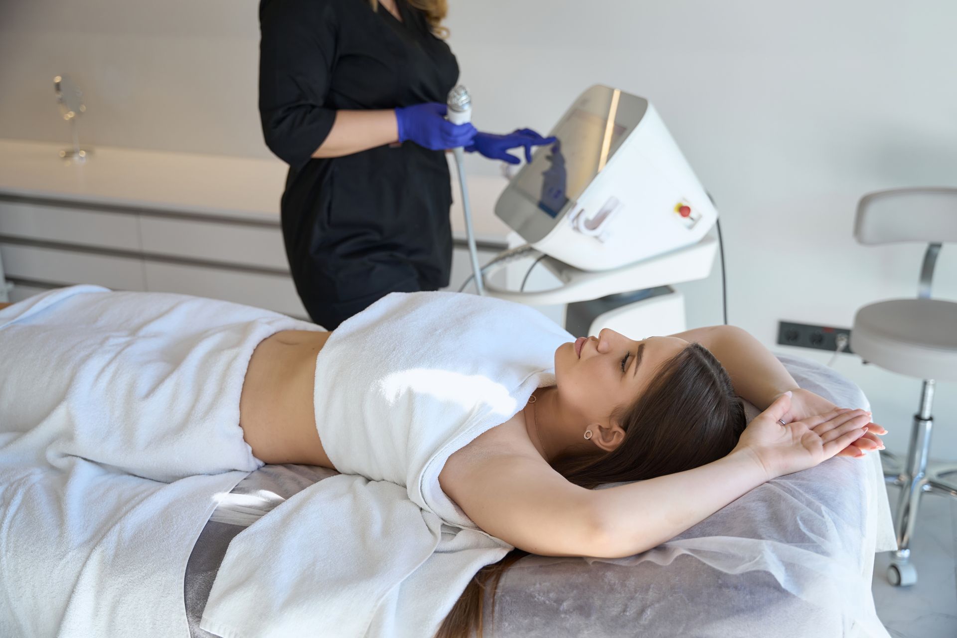 Woman receiving a facial treatment while lying on a treatment bed, with a technician using a device nearby