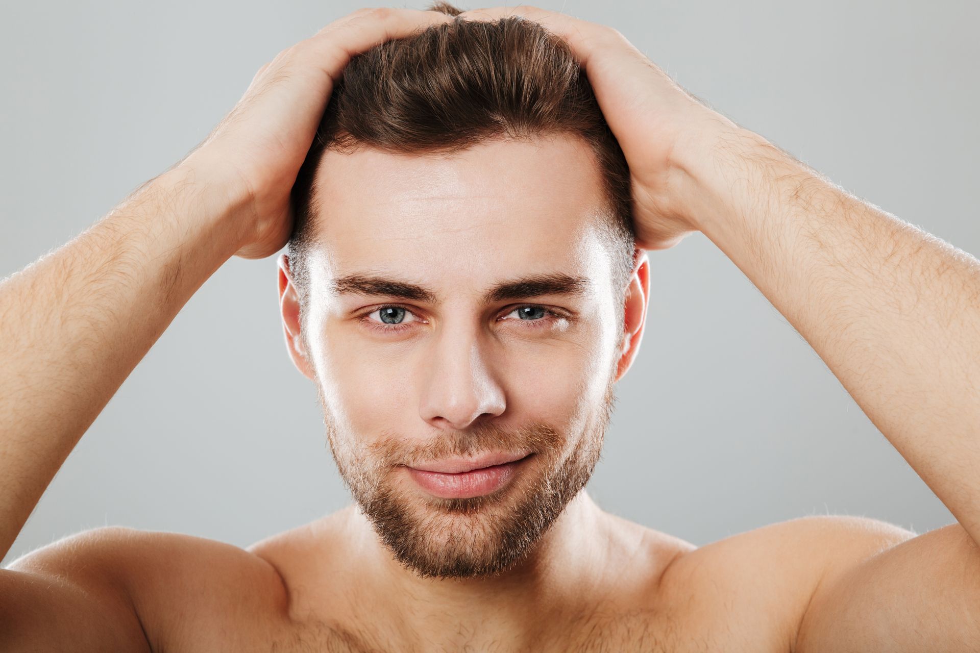 Man with hands in hair, smiling, bare-chested, against a gray backdrop.