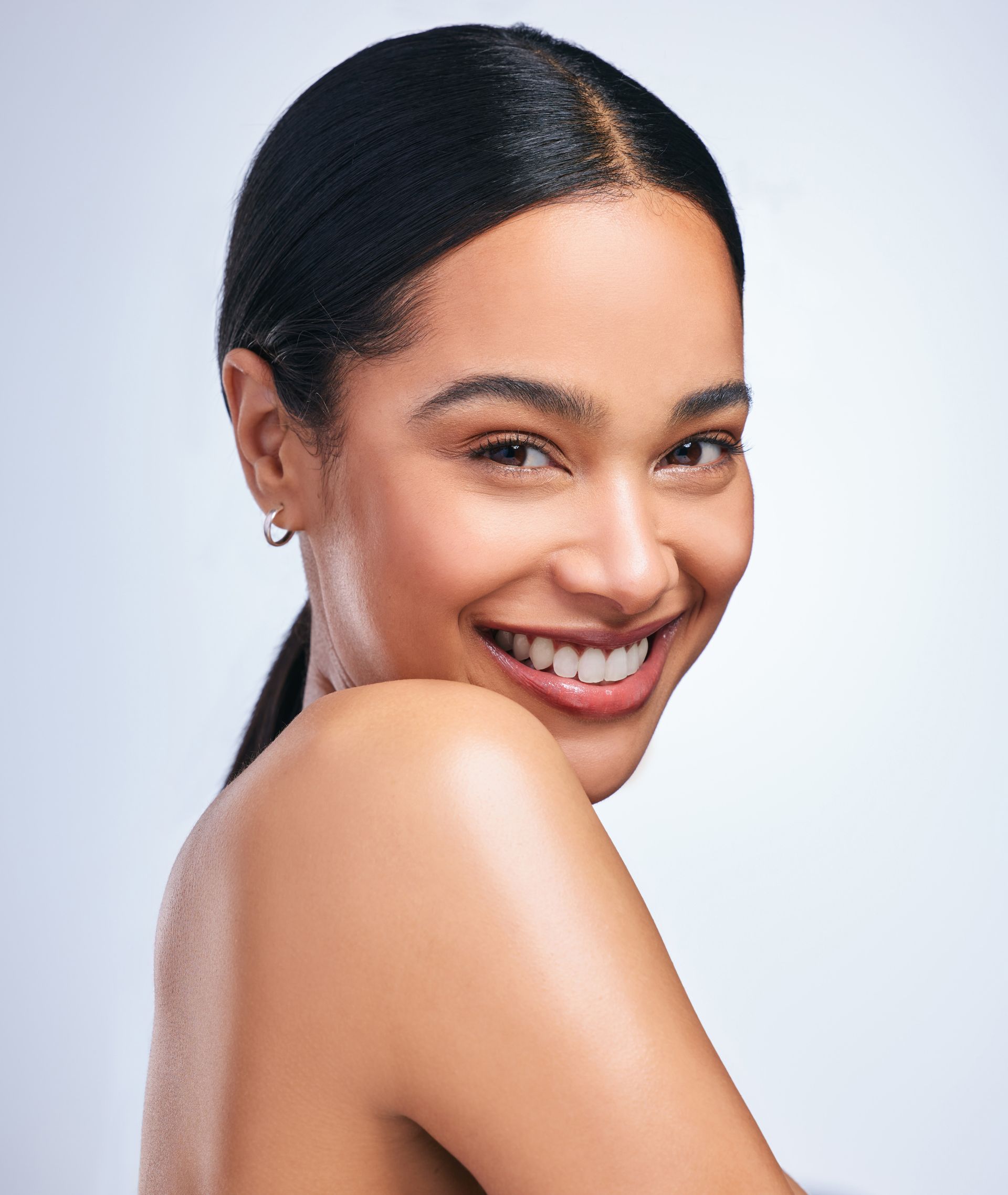 Smiling woman with dark hair in a sleek ponytail, bare shoulders, against a light gray background
