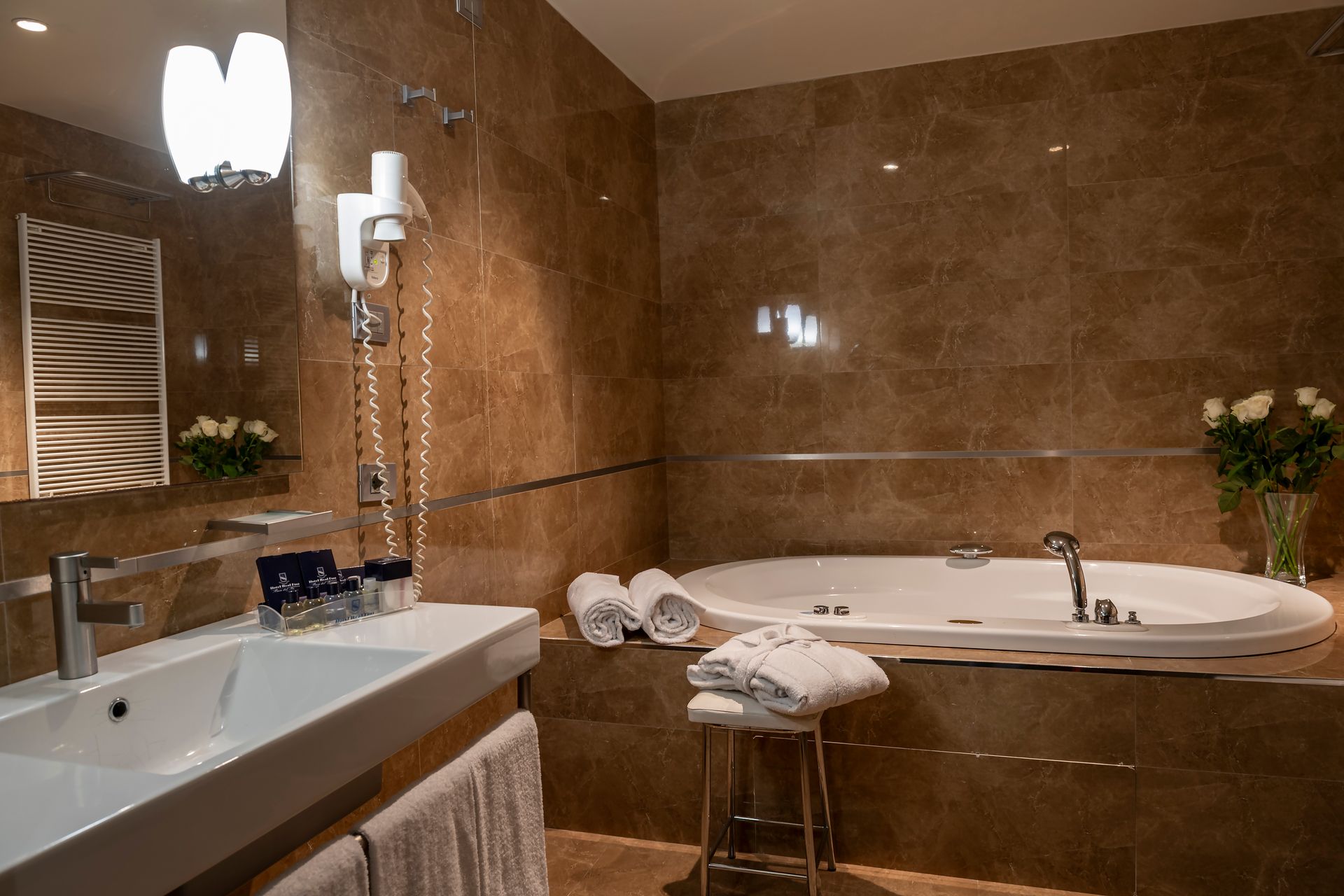 Salle de bains moderne avec murs carrelés marron, lavabo blanc, baignoire ovale, miroir et serviettes sur un tabouret.