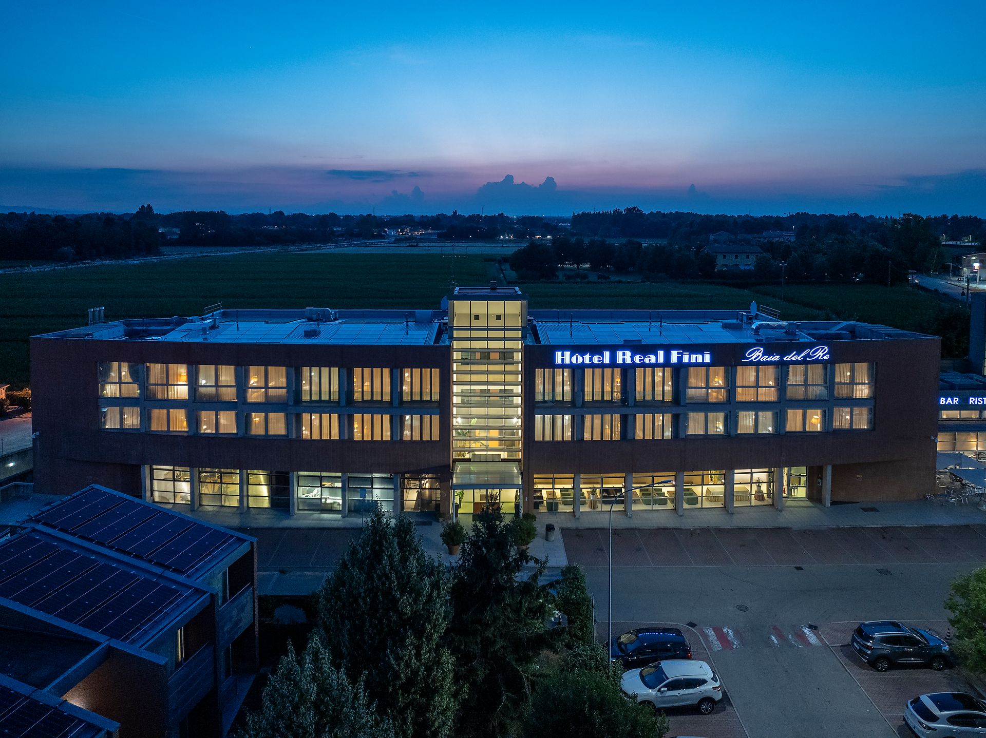 Hotel Real Fini Baia del Re au crépuscule, fenêtres éclairées et ciel rose-bleu au-dessus du parking