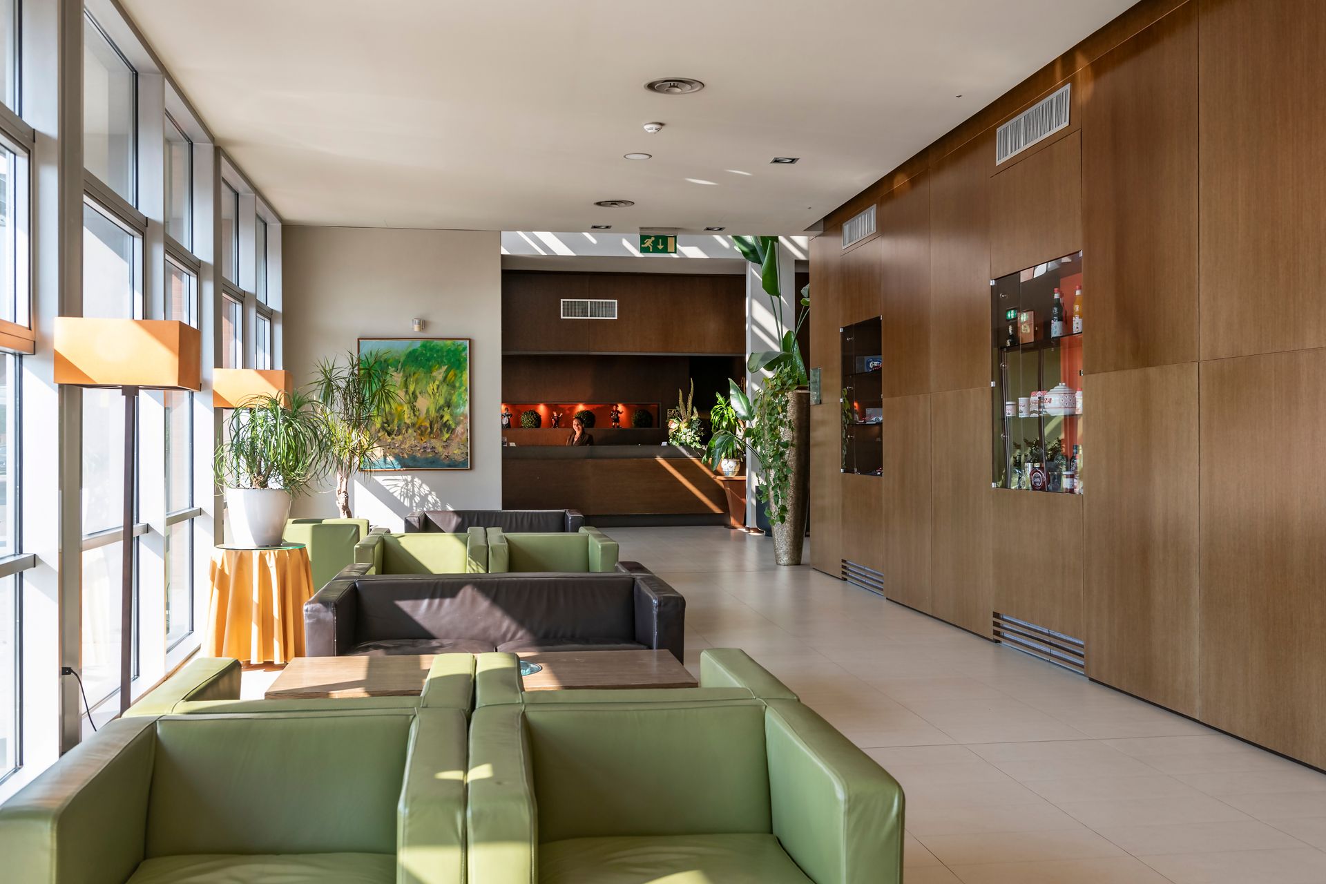 Salon d'hôtel moderne avec canapés verts, grandes fenêtres, mur lambrissé et espace réception à l'arrière.