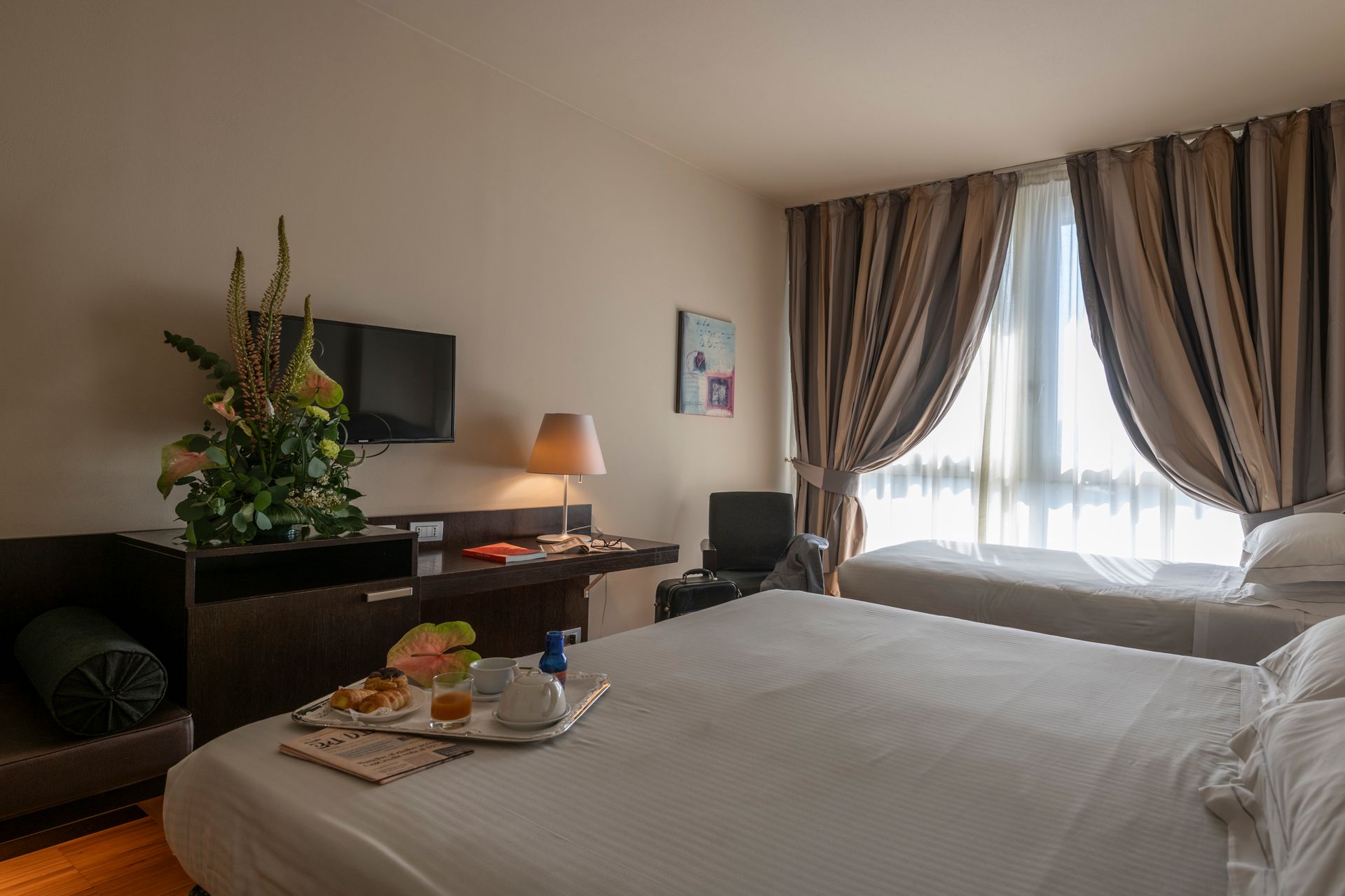Chambre d'hôtel avec un lit bien fait, un bureau, une télévision, une lampe et un plateau-repas près d'une fenêtre lumineuse