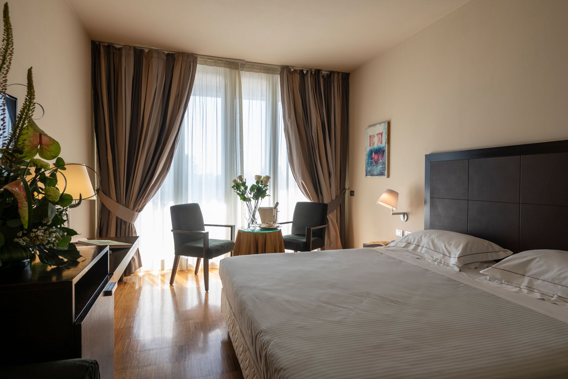 Chambre lumineuse avec un grand lit, des rideaux, deux chaises près de la fenêtre et une plante près de la porte.