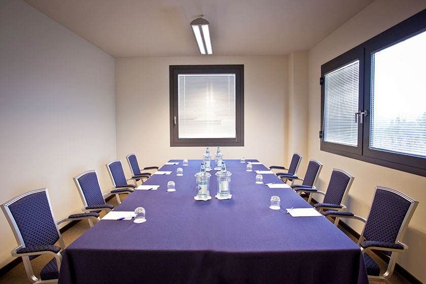 Salle de conférence avec une longue table, des chaises bleues et une nappe violette, éclairée par un plafonnier.