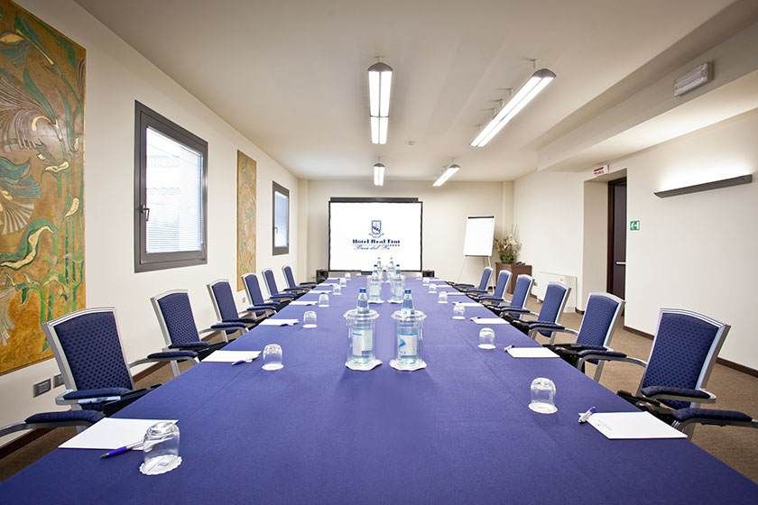 Salle de conférence avec une longue table bleue, des chaises noires et un écran de projection au fond.