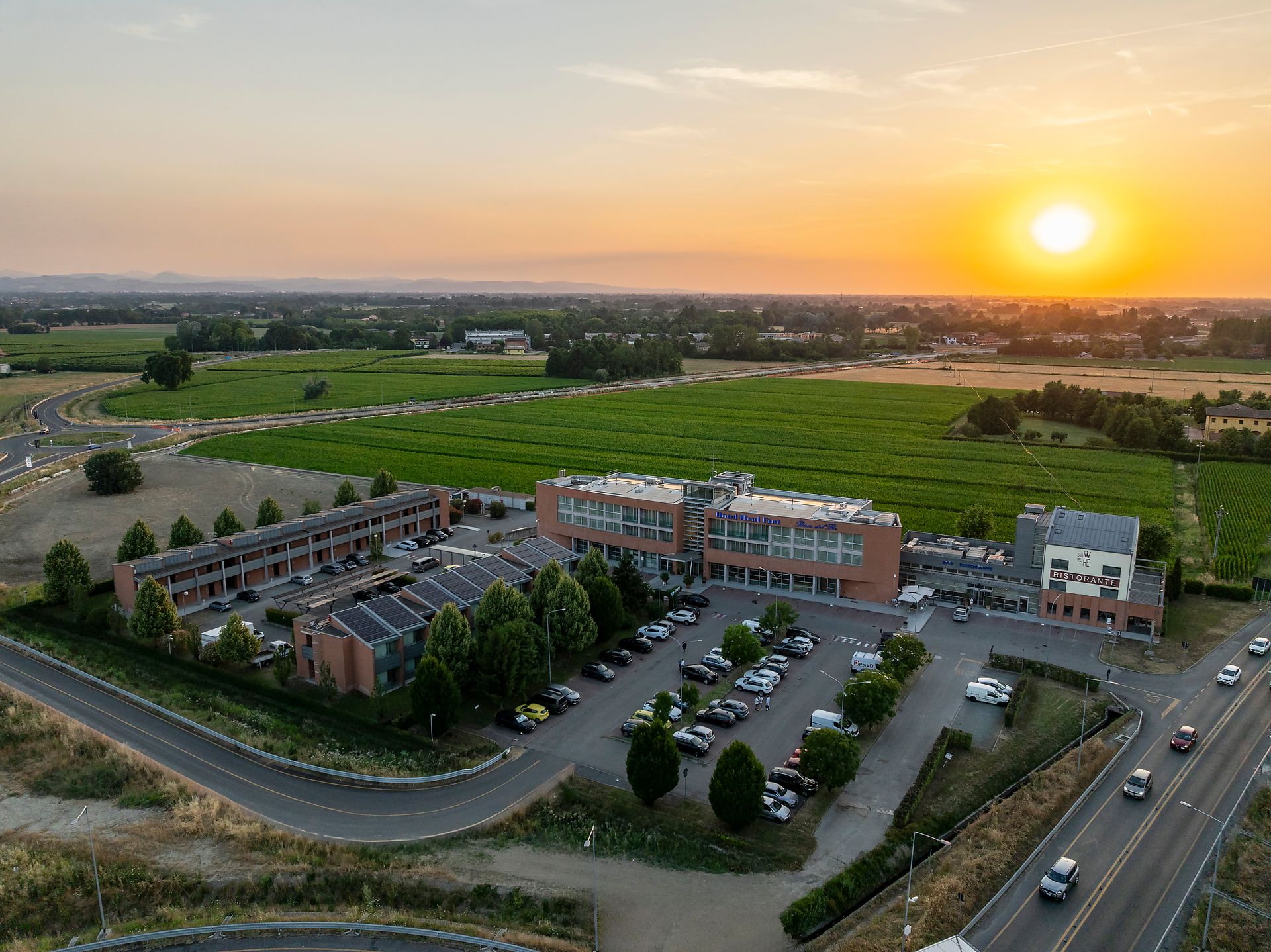 Vue aérienne d'Hotel Real Fini Baia del Re et d'un parking au coucher du soleil, avec des champs verdoyants à proximité.