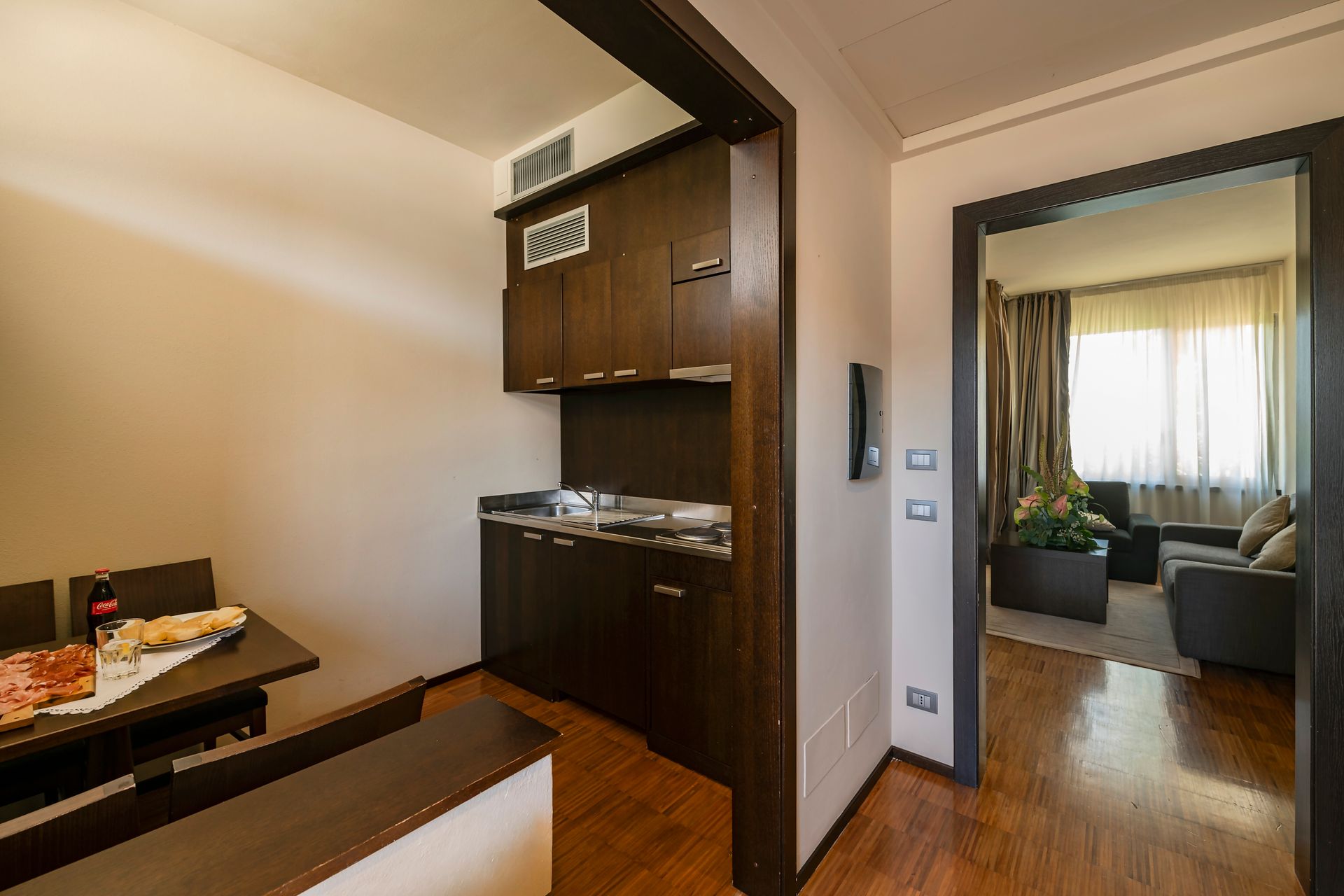 Petite chambre d'hôtel avec kitchenette, table et porte donnant sur un coin salon avec rideaux.