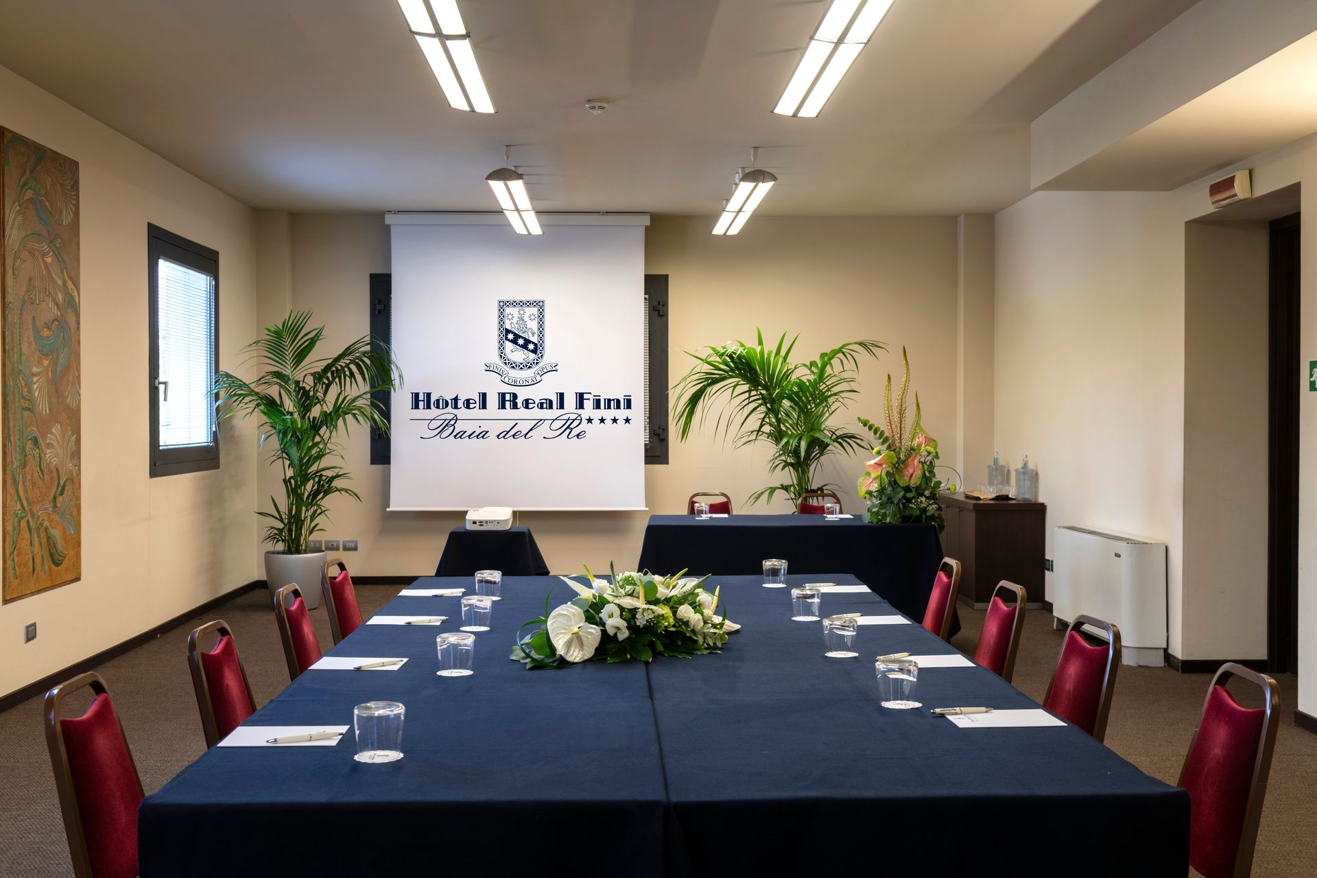 Salle de conférence avec une grande table, des chaises rouges, des plantes et un écran de présentation en façade