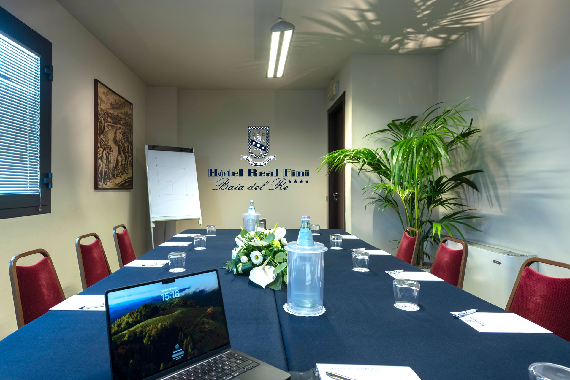 Salle de conférence avec une nappe bleue, des chaises rouges, des fleurs, une bouteille d'eau et un panneau mural au fond.