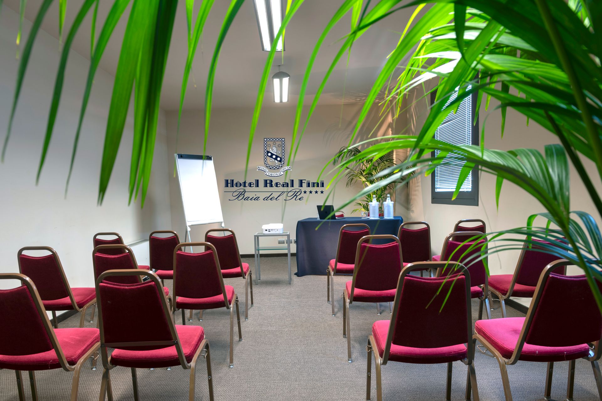 Salle de réunion vide avec des rangées de chaises bordeaux, un pupitre et un mur blanc encadré de plantes.