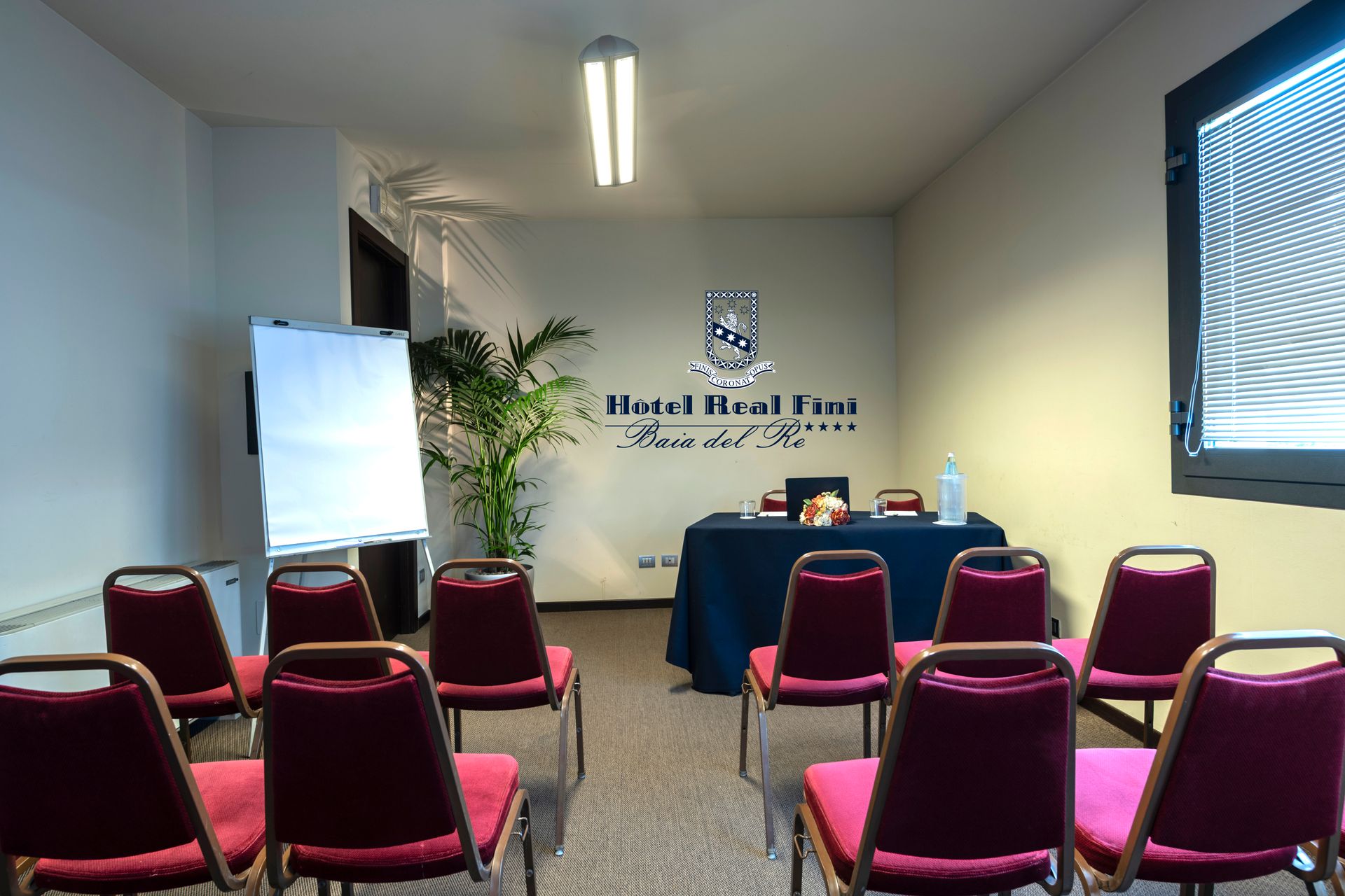 Petite salle de réunion avec des rangées de chaises rouges, un tableau de conférence et un logo mural au fond.