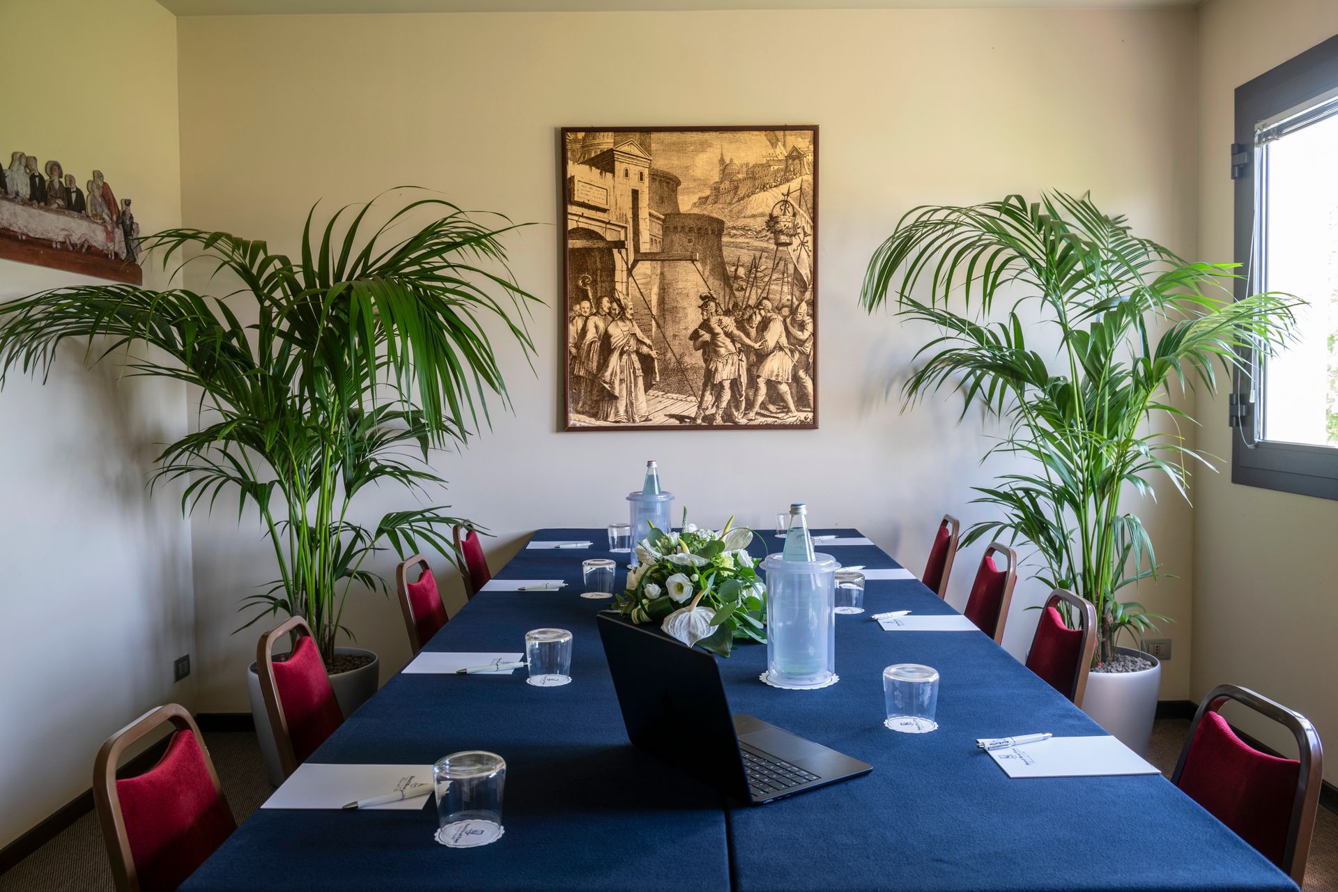 Salle de conférence avec une longue table bleue, des chaises rouges, des palmiers en pot et une photo encadrée au mur