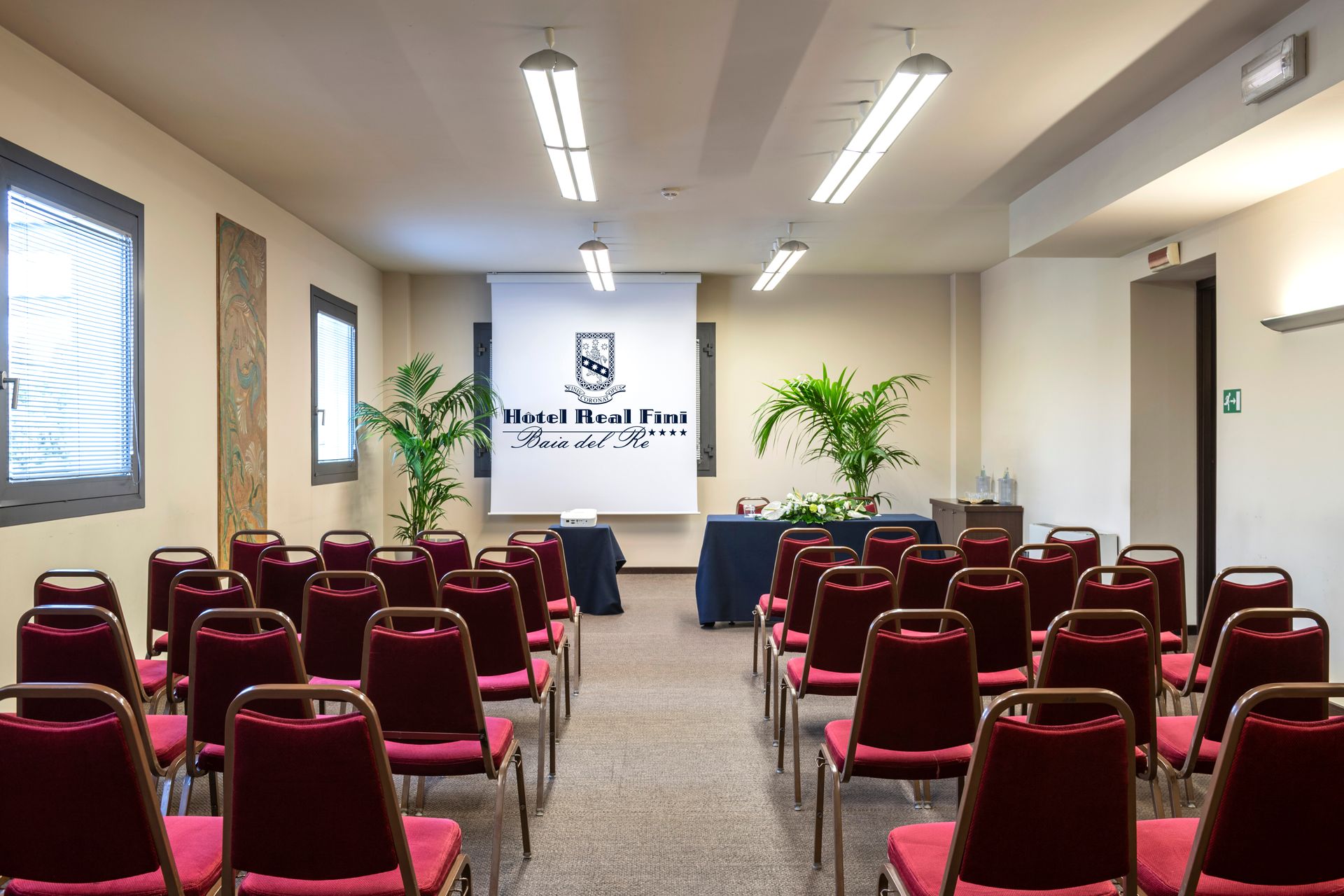 Salle de conférence vide avec des rangées de chaises rouges face à un écran de présentation et des plantes
