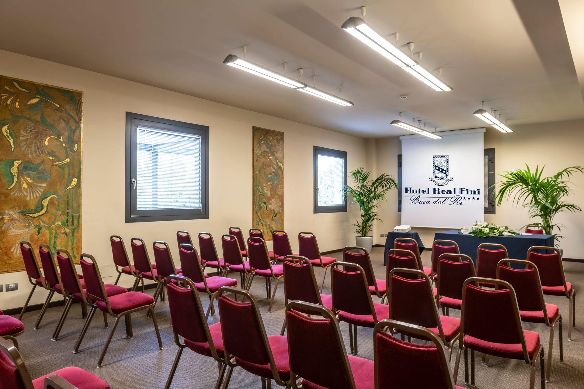 Salle de conférence avec des rangées de chaises rouges face à un écran de présentation et un podium.
