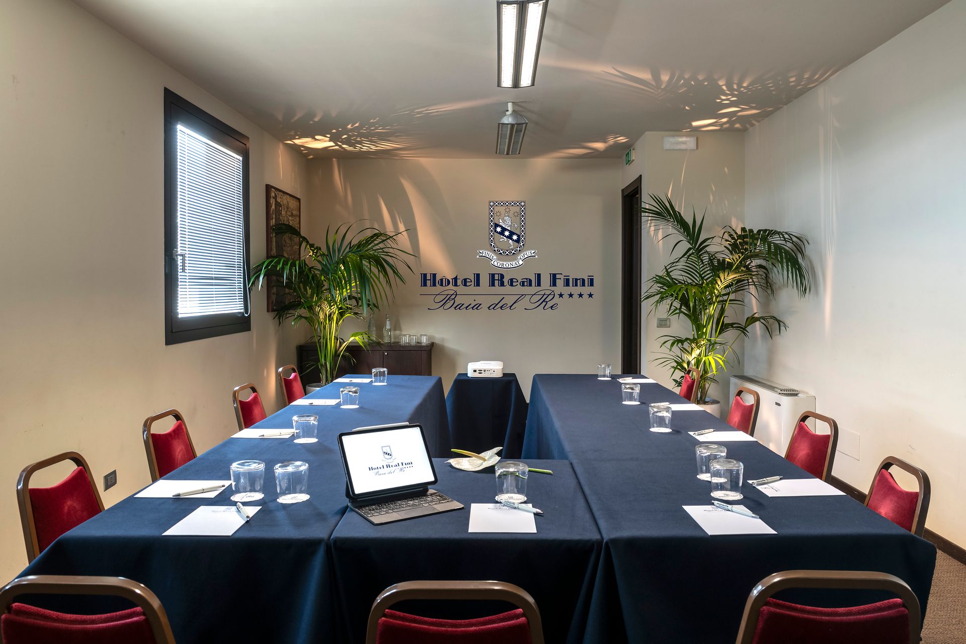 Salle de conférence avec une table en forme de U, des chaises rouges, des nappes bleues et un ordinateur portable au centre.