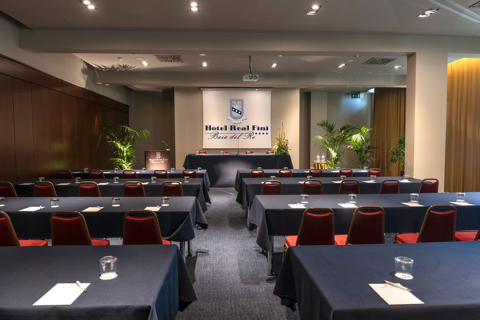 Salle de conférence avec des rangées de tables et de chaises face à une scène, éclairée par des spots encastrés.
