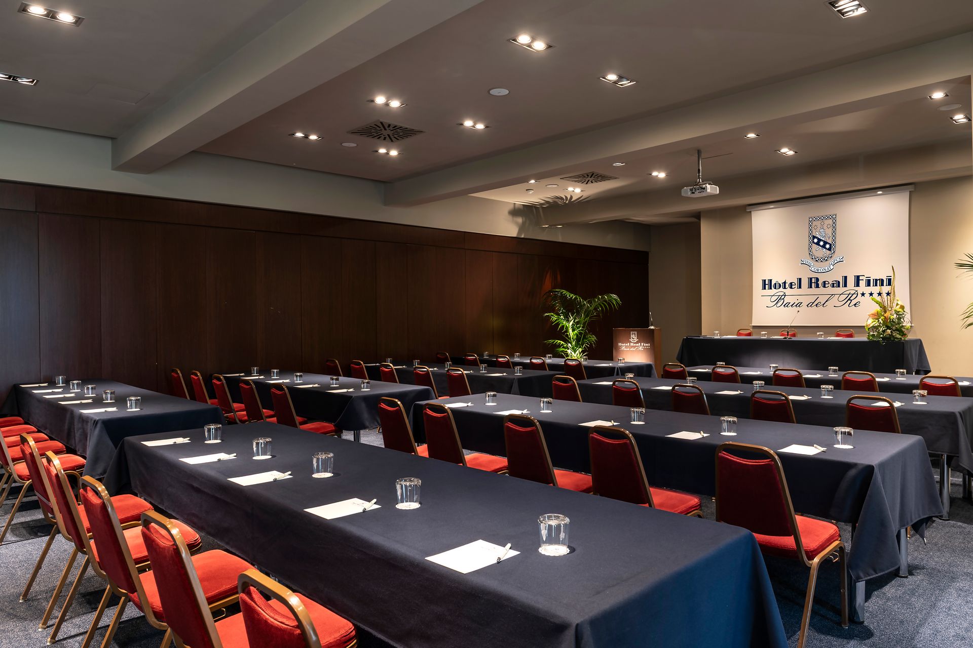 Salle de conférence avec des rangées de tables nappées de noir et des chaises rouges face à un écran de présentation
