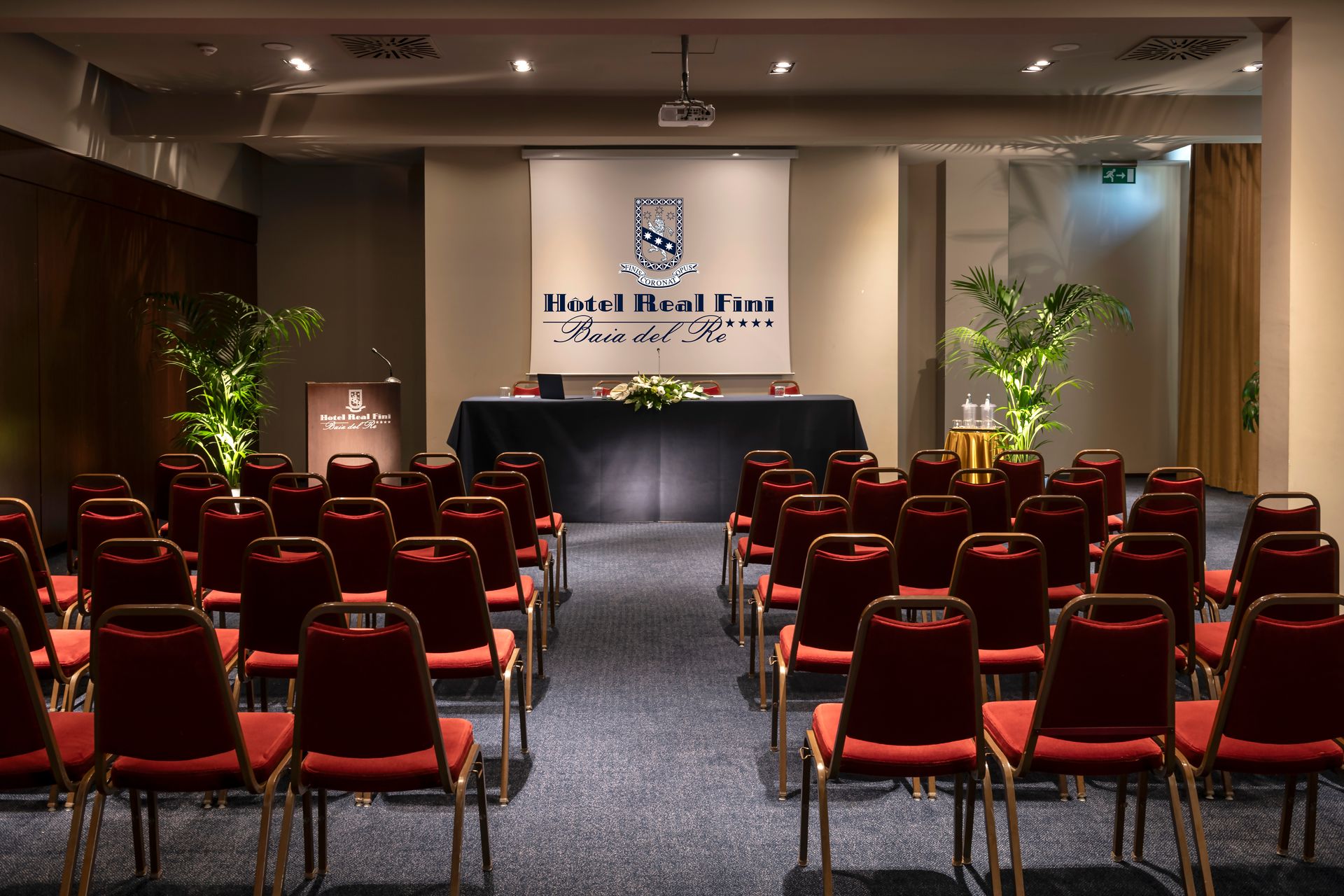 Salle de conférence avec des rangées de chaises rouges faisant face à une scène et un podium sous une banderole.