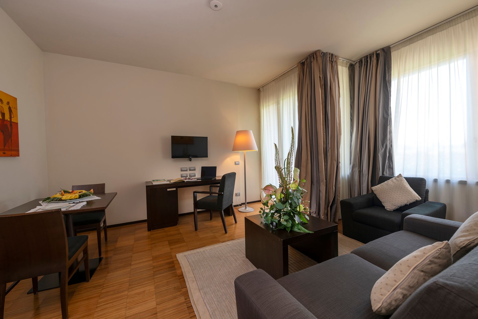 Salon moderne avec canapé, bureau, télévision, lampe et grande fenêtre avec rideaux.