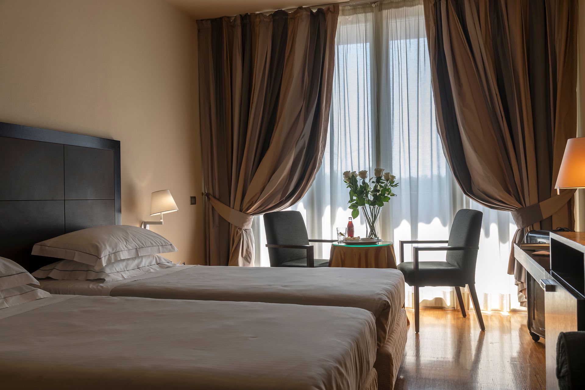 Chambre d'hôtel avec deux lits jumeaux, bureau, chaises et lumière du soleil filtrant à travers les rideaux.