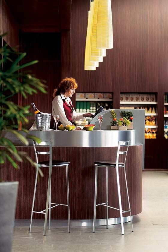 Une personne se tient derrière un bar incurvé dans un salon moderne avec des tabourets hauts et des suspensions lumineuses.
