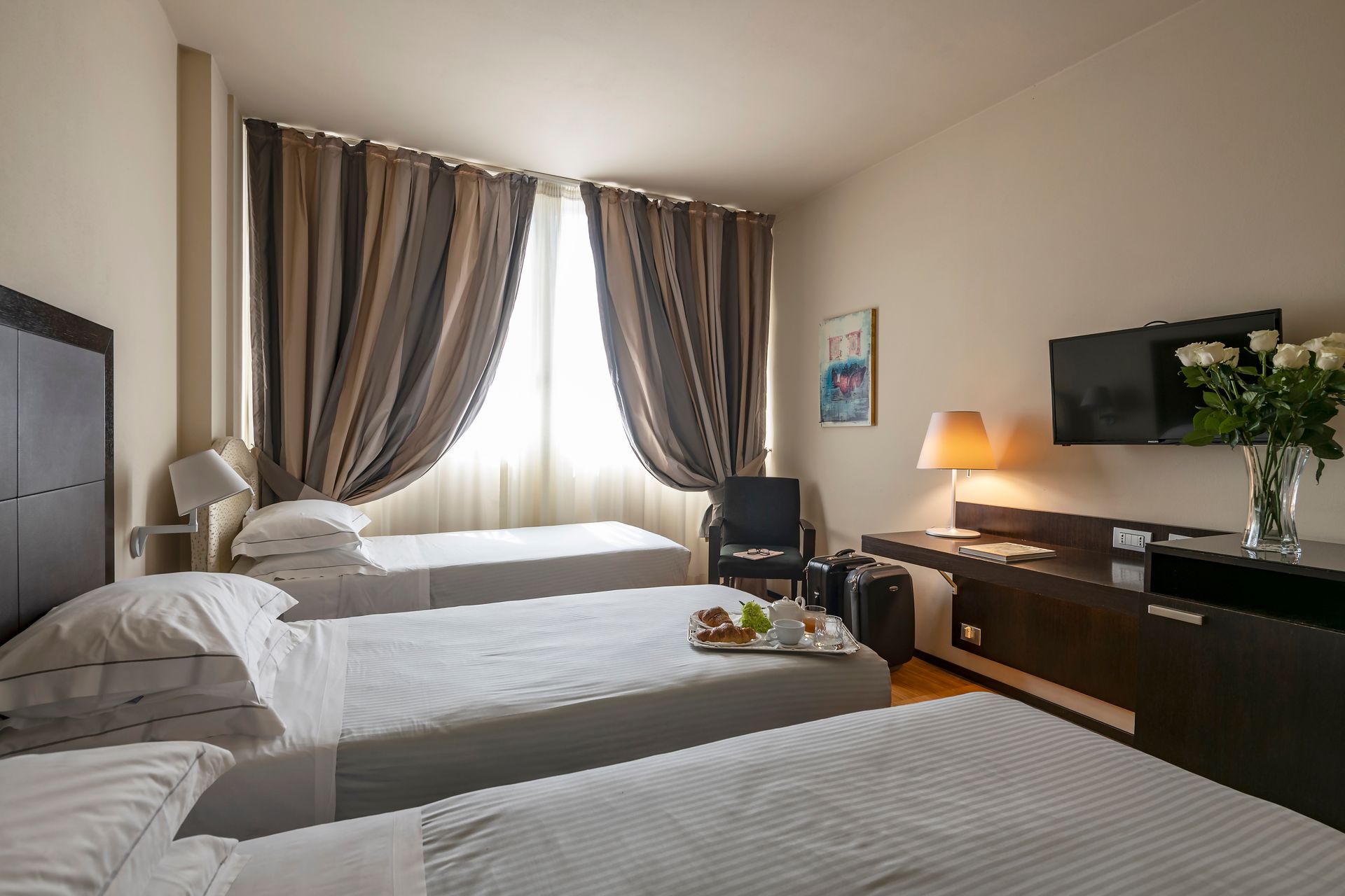 Petite chambre d'hôtel avec trois lits simples, fenêtre à rideaux, lampe de bureau, télévision et fleurs sur une commode.