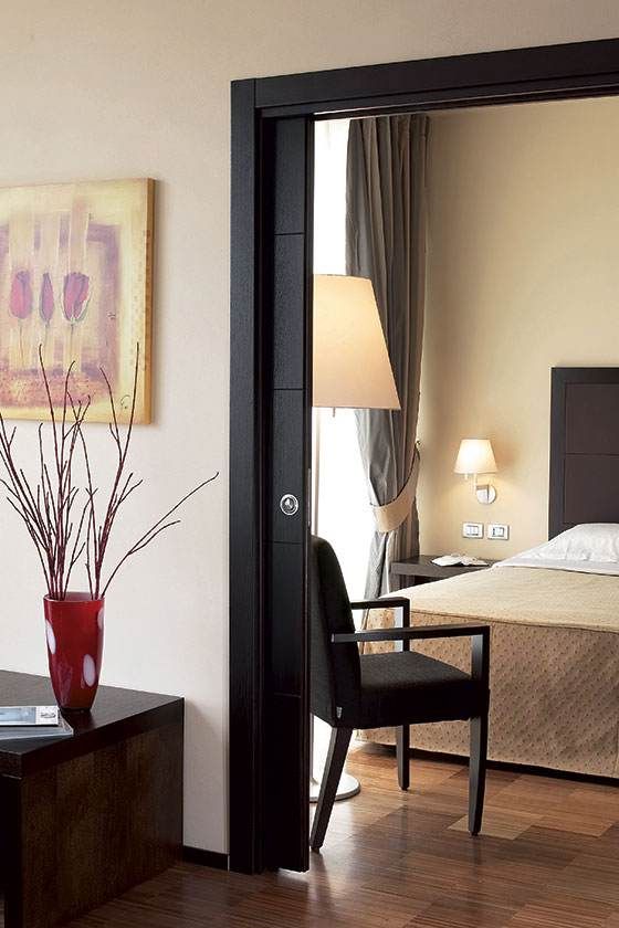Chambre vue à travers une porte sombre, avec un lit, une lampe, une chaise et un vase rouge au premier plan.
