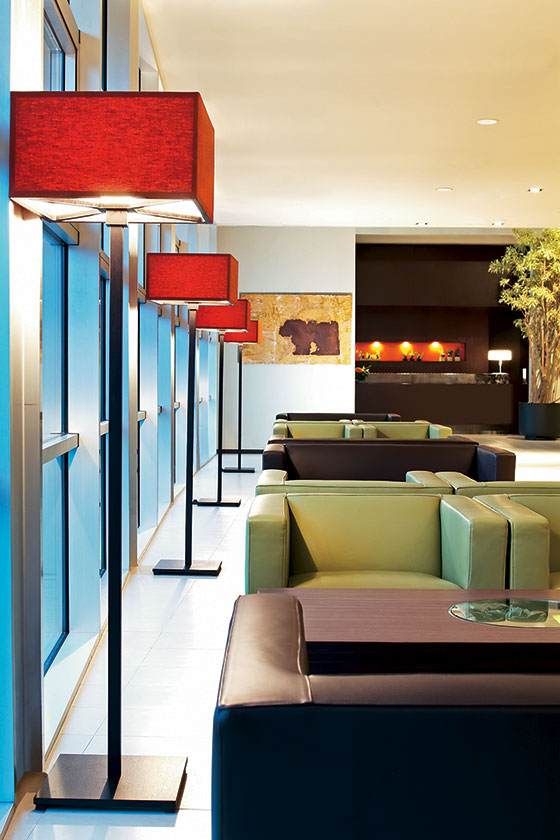 Salon moderne avec suspensions rouges, mur vitré bleu et sièges verts le long d'un couloir lumineux.