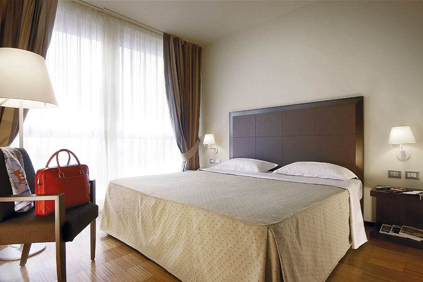 Chambre d'hôtel avec lit bien fait, rideaux aux fenêtres, lampes et un sac à main rouge sur une chaise