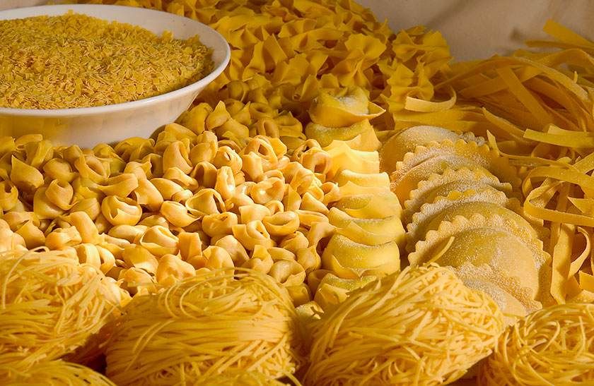 Assortiment de pâtes sèches jaunes de formes variées, disposées en tas, avec un bol de petites pâtes à l'arrière gauche.