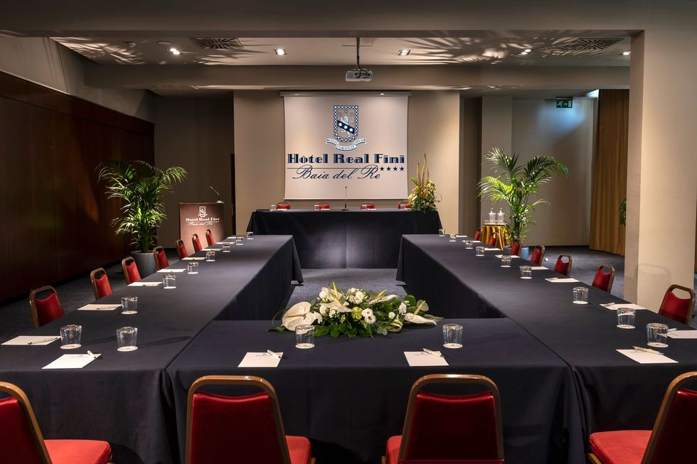 Conference room set for a meeting with U-shaped black-draped tables and red chairs, front podium and screen
