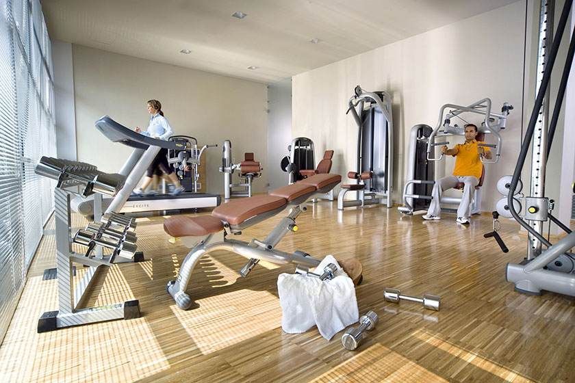 Salle de sport lumineuse avec tapis de course, appareils de musculation et banc, une personne s'entraînant en arrière-plan.