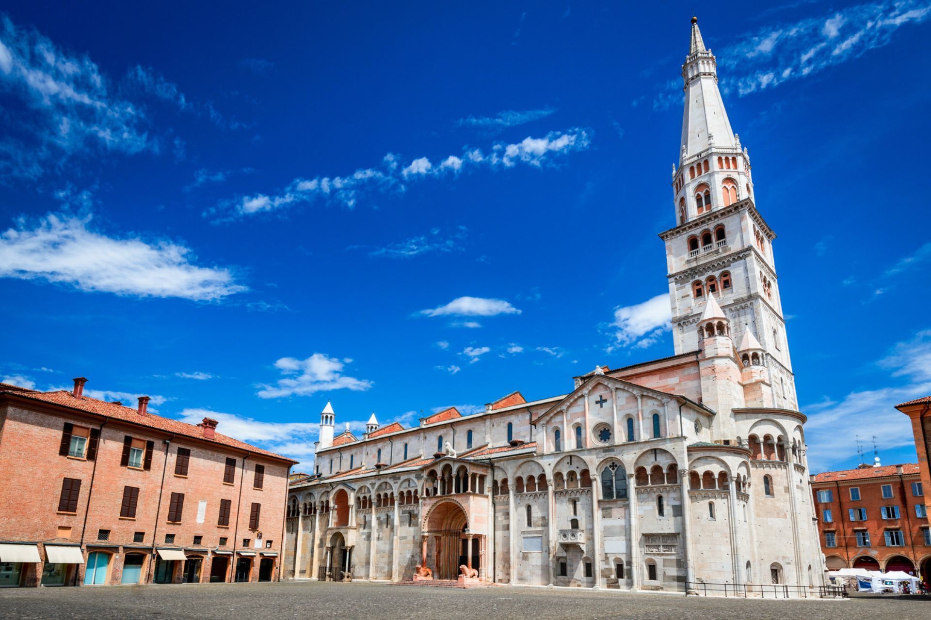 Storica cattedrale bianca e alto campanile in centro a Modena illuminata dal sole, sotto un cielo azzurro intenso.