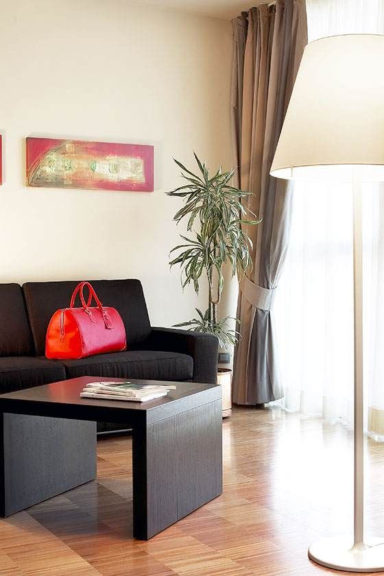 Luminoso soggiorno con divano nero, borsa rossa, tavolino da caffè, pianta e lampada da terra vicino a una finestra.