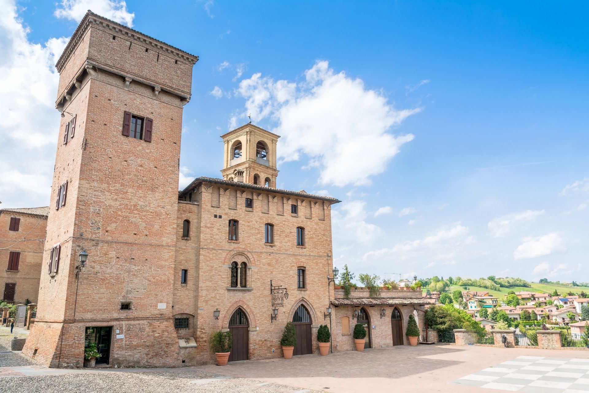Piazza di Castelvetro di Modena con la torre medievale in mattoni, la pavimentazione a scacchiera e le colline sullo sfondo