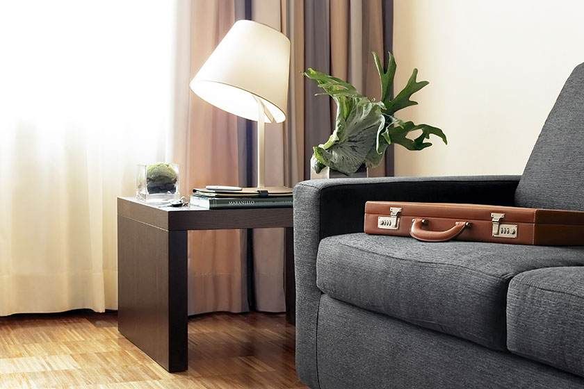 Salon moderne avec canapé gris, table d'appoint, lampe, rideaux et valise sur le canapé.