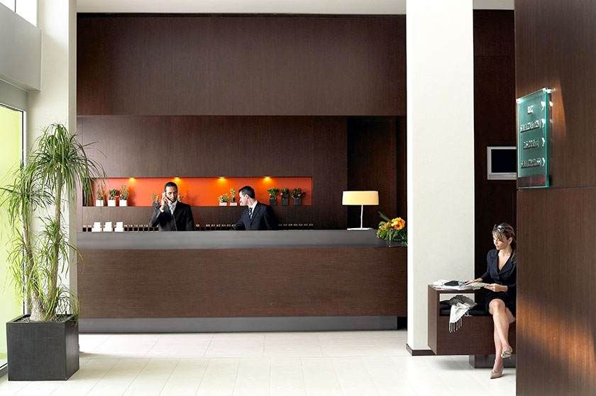 Hall d'hôtel moderne avec réception, deux employés et un client assis près d'une lampe.