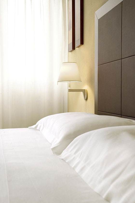 Lit blanc avec oreillers à côté d'une applique murale et d'une fenêtre dans une chambre d'hôtel à l'éclairage tamisé