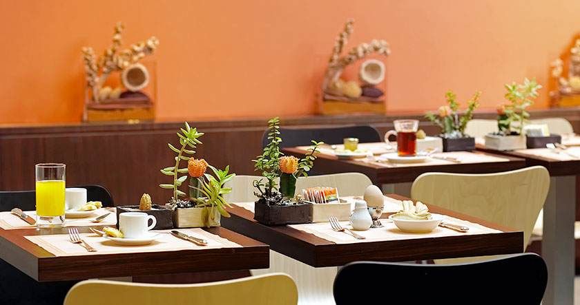 Salle à manger chaleureuse avec tables en bois, couverts blancs, plantes en pot et murs orange.