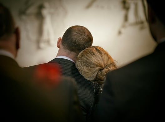 Man and woman embrace, woman resting head on man's back, others in suits near them.