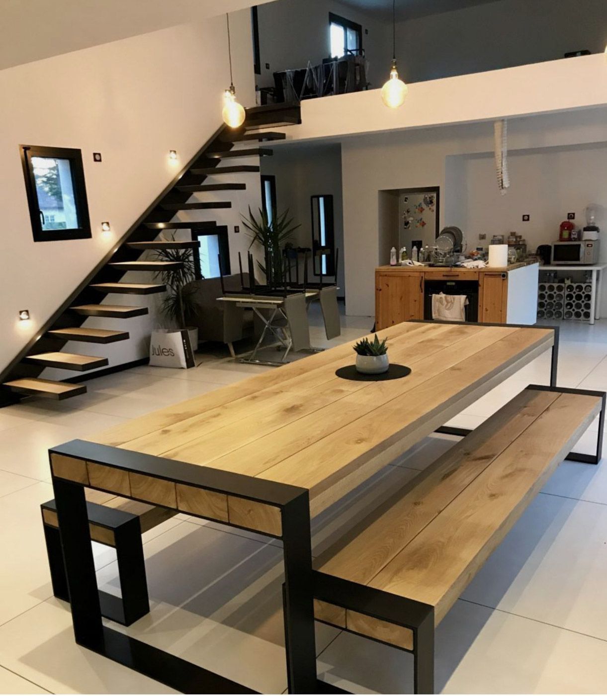 Zona pranzo in stile loft moderno con un lungo tavolo in legno, panche e una scala che conduce al piano superiore.
