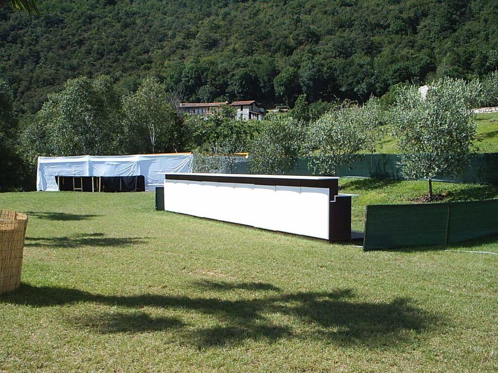 Terrazza erbosa esterna con fioriere basse bianche e scure, alberi e una collina boscosa sullo sfondo.