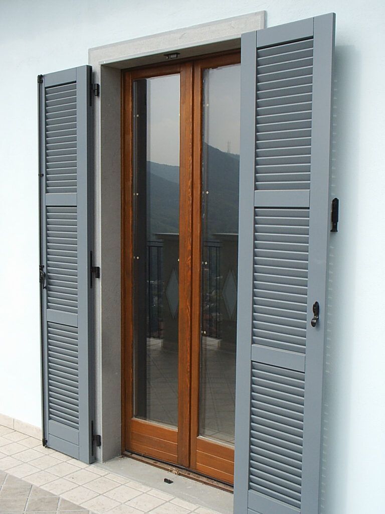 Persiane metalliche grigie che fiancheggiano una porta-finestra in vetro con telaio in legno che delimita il balcone.