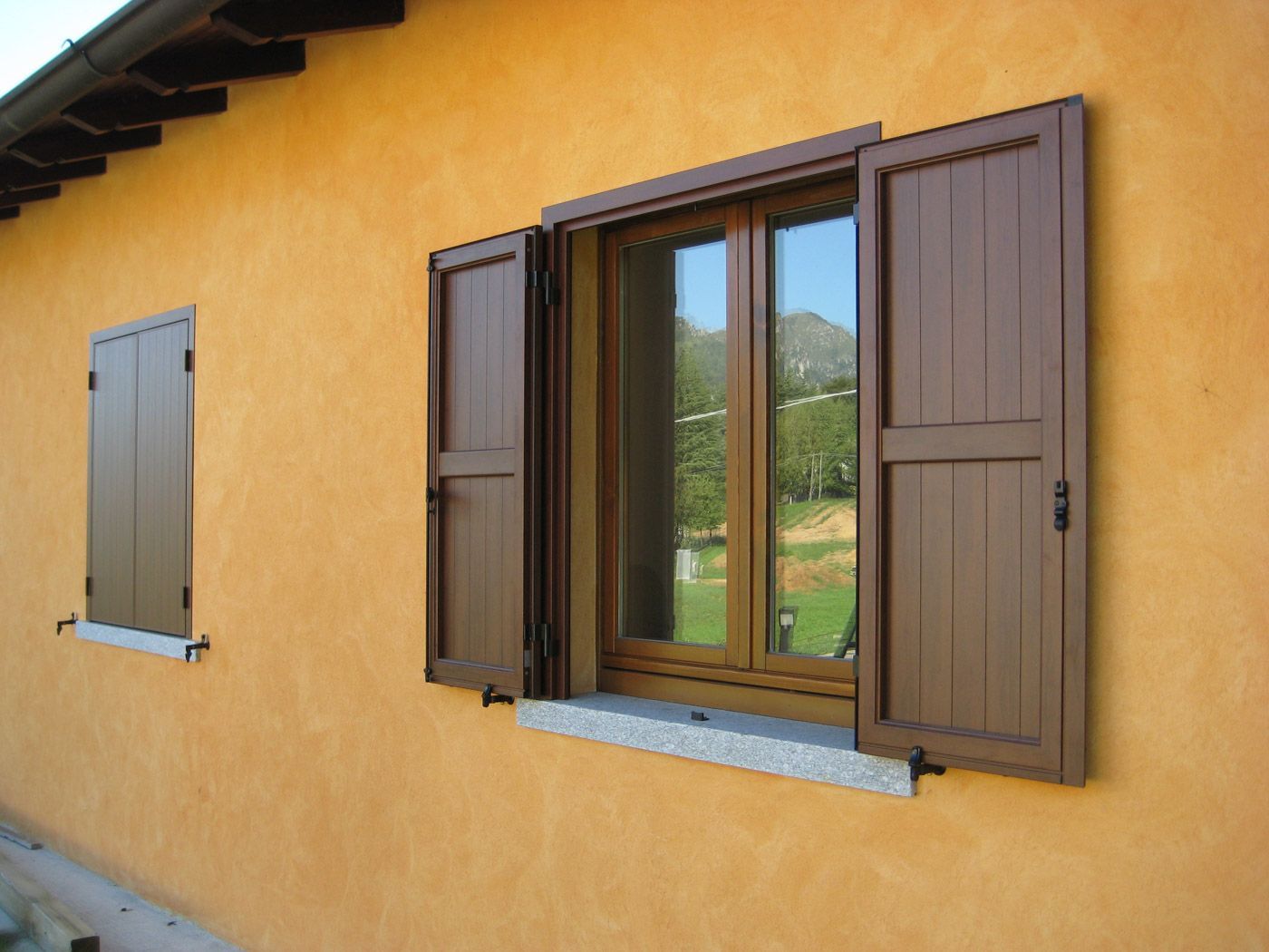 Muro di casa in stucco giallo con due finestre, una aperta con persiane marroni e vista sul verde.